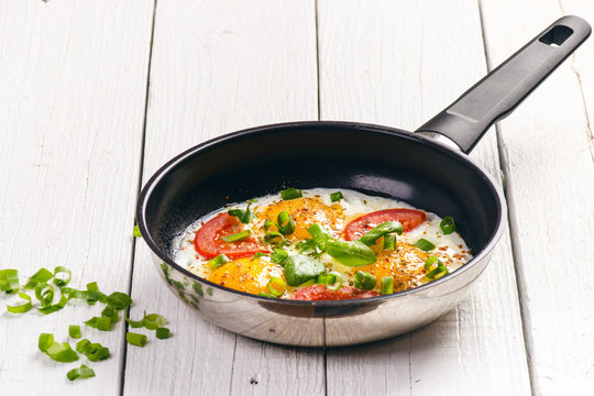 Pan of fried eggs with tomatoes, cheese, spring onion, herbs on a white table. White wooden table. Concept of food. Breakfast time. Copy space. 