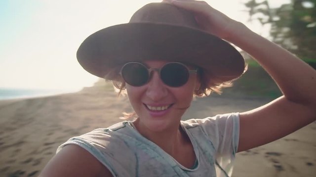 A young woman with hat in the sunlight on the ocean beach smiles on camera enjoys new adventures, vacation and travel. Portrait of a tourist in a beach with palm trees on background