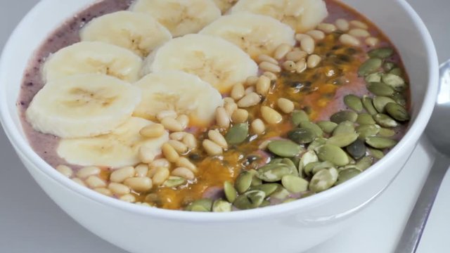 Hearty serving of smoothie topped with nuts, seeds, passionfruit and banana slices in white bowl. Tilt up shot. Concept of healthy food and lifestyle