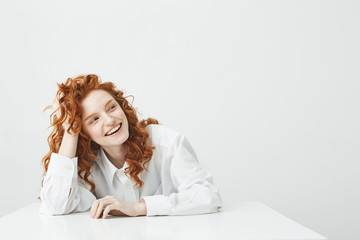 Naklejka premium Cheerful pretty young girl with foxy hair smiling laughing sitting at table over white background.