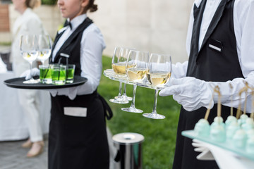 Midsection of professional waiters in uniform serving wine and snacks during buffet catering party, festive event or wedding. Full glasses of champagne on tray.