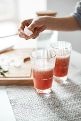 Close up of girl's hands putting ice pieces in glasses with grapefruit detox healthy smoothie.