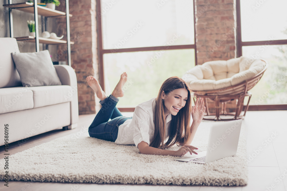 Wall mural Hello there! Young cheerful lady is waving to the camera while having video call lying on the carpet at home, so happy and excited