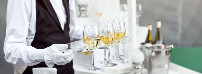Midsection of professional waiter in uniform serving wine during buffet catering party, festive event or wedding. Full glasses of champagne on tray.