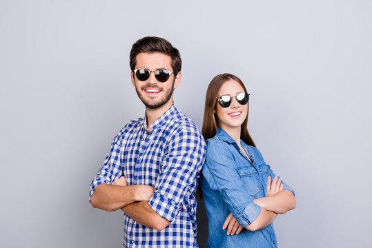 Two cheerful young lovers are looking in the camera, wearing stylish sunglasses and smile, in casual shirts, with crossed hands on the pure background