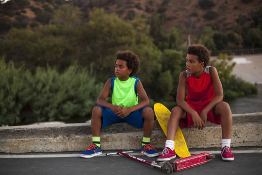 Two brothers with skateboard and push scooter watching from kerb