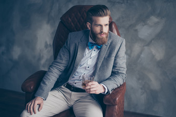 Successful young red bearded elegant businessman in suit with perfect hairstyle is drinking whiskey indoors, relaxing, sits on leather arm chair, looks harsh and virile - obrazy, fototapety, plakaty
