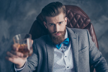 Cheers, ladies and gentlemen! Stylish elegant red bearded aristocrat in suit and bowtie holds glass with brandy, relaxing, sits on leather brown arm chair indoors - obrazy, fototapety, plakaty