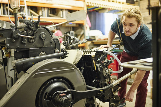 Young man operating old printing machine in traditional print workshop
