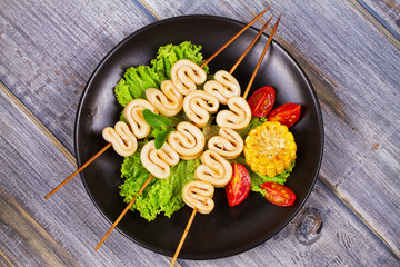 Grilled squid rings on skewers and vegetables on black plate. View from above, top studio shot