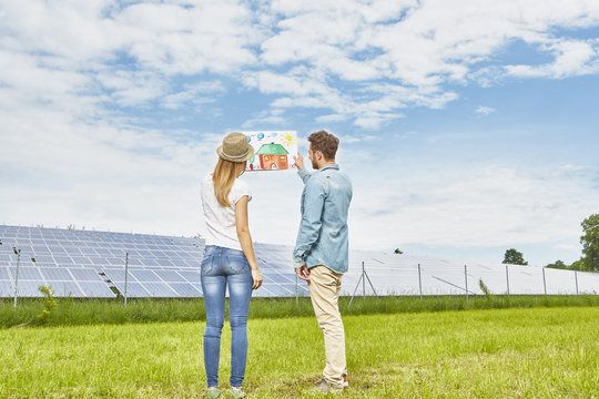 Young couple standing in field, looking at child's drawing of house, next to solar farm