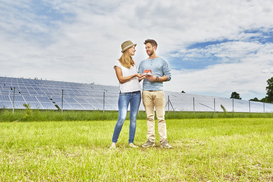 Young couple standing in field, holding small model of house, next to solar farm