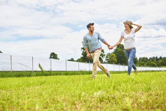 Young couple holding hands, running through field, next to solar farm