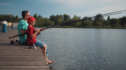 Side view of man with little boy sitting on wooden pier fishing on background of lake.