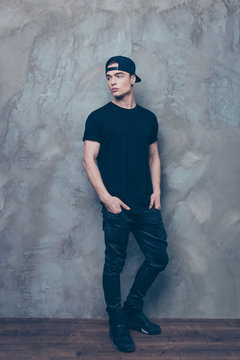 Full size portrait of attractive young man in cap and black cool outfit is on the grey background, his hands are in pockets, he looks away