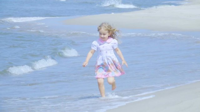 Adorable little girl  in a dress runs  jumping on the beach.  Child enjoys playing on the seashore running on the water with pleasure. splashing in sea waves.Slow Motion