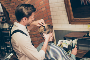 Back view of a bearded stylish barber shop client. He is getting his perfect trendy haircut from a classy dressed stylist, reading the magazine and waiting for result