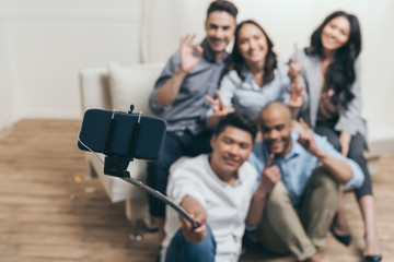 young smiling multiethnic friends taking selfie with monopod and smartphone at home