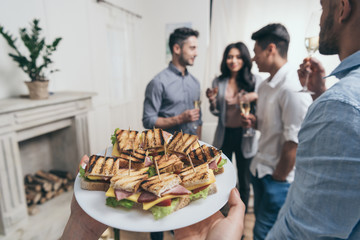 Cropped shot of person holding plate with tasty sandwiches while smiling young friends drinking...