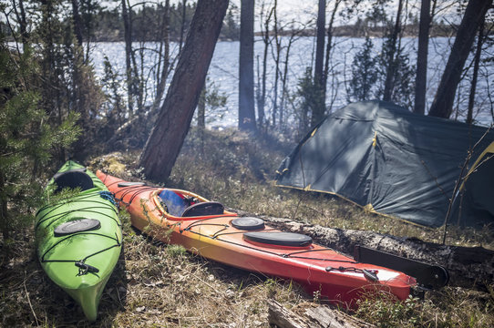 Two kayaks are in the spring forest near the camp, the sun and the smoke from the fire, near the lake and tents.