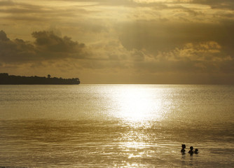 Obraz premium Small silhouettes of a man, woman and baby bathing in the ocean on a golden sunset with tropical land silhouette on the background