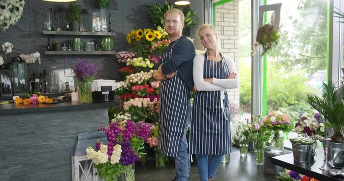 SIde view of young cheerful coworkers in striped aprons standing back to back with hands crossed posing in floral shop.