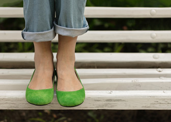 Female legs in blue jeans and green shoes on a white bench.