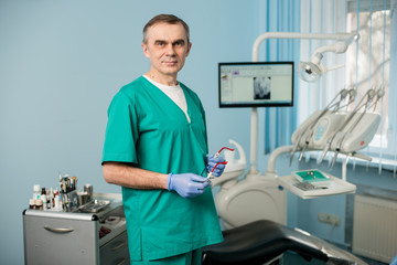 Portrait of handsome male dentist at the morden dental office. Doctor wearing green uniform, blue gloves, holding glasses and looking to the camera. Dentistry