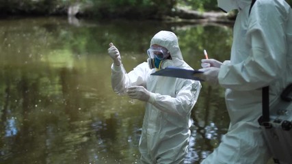 Biochemist in suit taking sample of water in test tube while investigating nature with coworkers.
