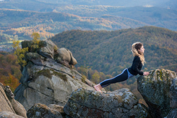 Young fit female is practicing yoga and doing asana Urdhva Mukha Shvanasana on the top of the mountain. Autumn forests, rocks and hills on the background