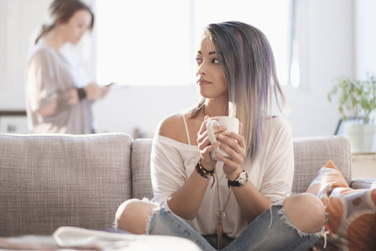 Young woman sitting on sofa and holding mug, other woman using mobile phone