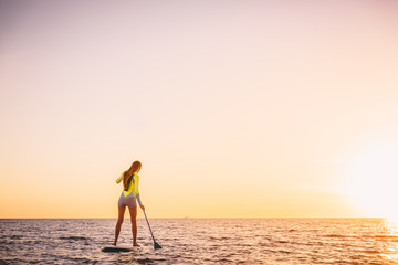 Slim sporty woman in ocean on stand up paddle surfboard with beautiful sunset colors