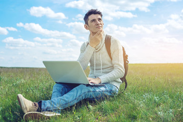 Young man sitting on a green meadow with laptop wireless on the background of blue cloudy sky
