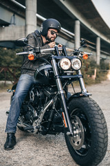 Fototapeta na wymiar Young brutal man in a black jacket and glasses on motorcycle. Male biker wears a black helmet.