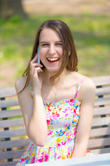 Fototapeta premium Portrait of young beautiful woman with long hair in summer park, girl wearing flower short dress is using a smartphone while sitting on a bench
