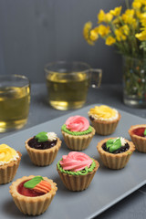 Colorful cupcakes on grey plate and table. Cakes with cream and jam. Cup of green tea and yellow flowers on a background