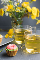 Colorful cupcakes on grey plate and table. Cakes with cream and jam. Cup of green tea and yellow flowers on a background