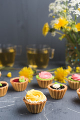 Colorful cupcakes on grey plate and table. Cakes with cream and jam. Cup of green tea and yellow flowers on a background