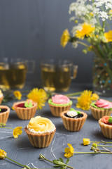 Colorful cupcakes on grey plate and table. Cakes with cream and jam. Cup of green tea and yellow flowers on a background
