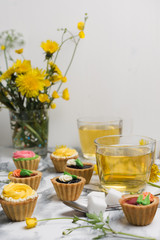 Colorful cupcakes on grey marble table. Cakes with cream and jam. Cup of green tea and yellow flowers on a background