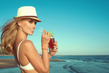 Portrait of young glam smiling blond lady in white swimming bra and panama standing at the seaside drinking cocktail through a straw and looking aside. Her hair blown by the wind. Copy space