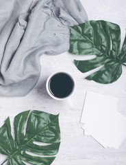 Black coffee and big green tropical leaves next to a grey scarf and white pieces of paper on grey concrete background. Flat lay. Top view. Copy space
