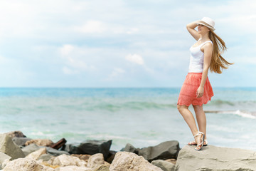 Naklejka premium young pretty woman with white hat and red skirt is staing on stone at seashore under gray sky with strong sea background