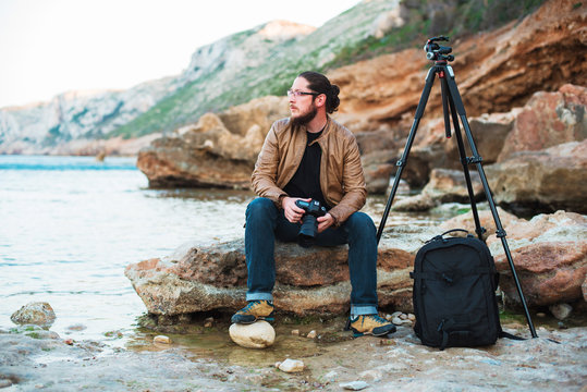 Young stylish photographer sitting on rock and looking at sea with camera in hands