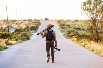 Young photographer walking on desert road with tripod on his shoulder and camera in hand - obrazy, fototapety, plakaty