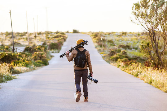 Young photographer walking on desert road with tripod on his shoulder and camera in hand