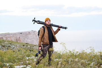 Fototapeta premium Young photographer walking in the nature with tripod on his shoulder and camera in hand