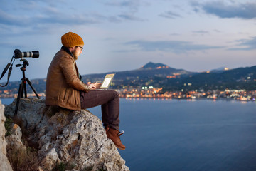 Young attractive photographer sitting on rock and working on laptop with view of sea and mountains