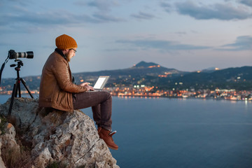 Young attractive photographer sitting on rock and working on laptop with view of sea and mountains