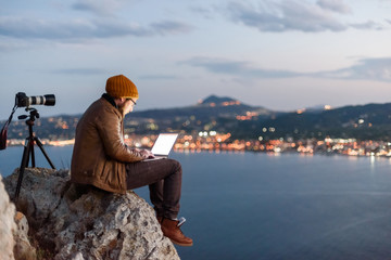 Fototapeta premium Young attractive photographer sitting on rock and working on laptop with view of sea and mountains